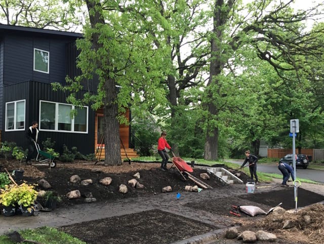 Complete Ecological Landscaping Services - Light Dark Landscape 3 Four landscape crew members installing new boulders and plants into fresh dirt in front of a modern black and natural wood house on an urban street.