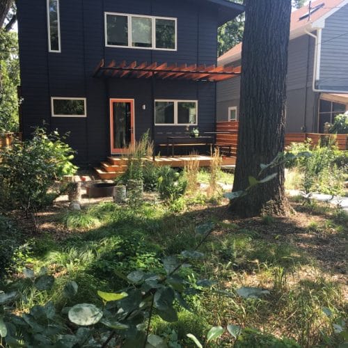 A fenced backyard with a large oak tree in front of a modern black house with a natural wood patio leading to a rustic fire pit.A fenced backyard with a large oak tree in front of a modern black house with a natural wood patio leading to a rustic fire pit.