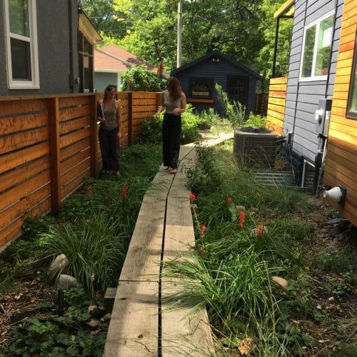 Two women talking on a boardwalk leading through the narrow side of a modern black and natural wood home. The boardwalk is suspended over a rain garden with blooming red flowers.