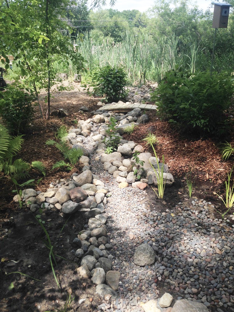 A rocky dry steam with a bird feeder, grasses, and a small bridge over the creek.