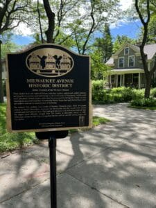 A black and gold illustrated sign explains the history of the Milwaukee Avenue Historic District and depicts an illustration of three Victorian era row houses.