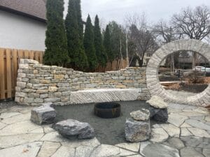 A flagstone patio, boulder-lined gravel fire pit, undulating dry-stacked wall and dramatic moon gate are key elements of a newly constructed outdoor kitchen and beer garden, situated on a private property on Kenwood Avenue in Minneapolis.
