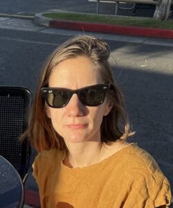 A woman with light brown hair and sunglasses facing the camera wearing an ochre colored shirt for a portriat about her.