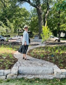 A woman wearing a hat, blue shirt, and black skirt standing on a cobbled stone path with new plants on either side on a tree-lined street for a portrait about her.