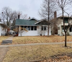 A quaint, cabin-like white house with green trim sits on a small lot with a bare lawn, foundation shrubs