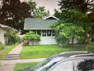 A quaint, cabin-like white house with green trim sits on a small lot with a bare lawn, foundation shrubs and a single Tamarack tree.