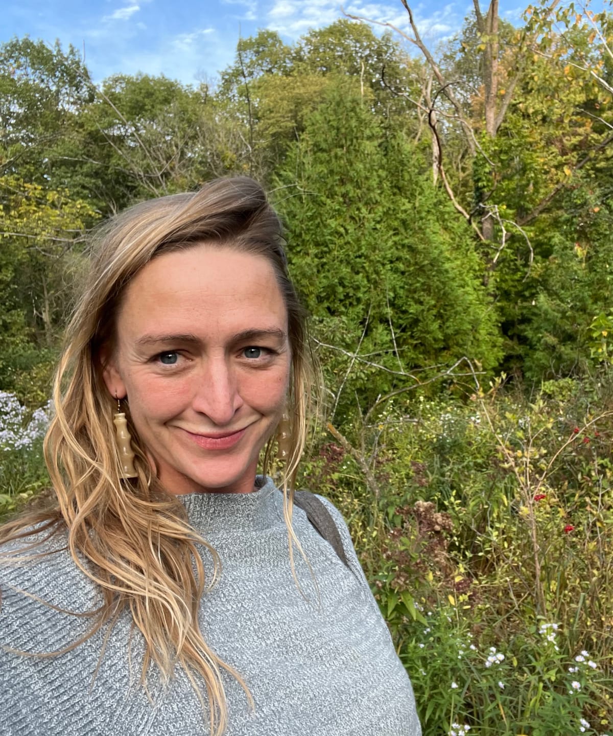 Our Landscaping Team - Light Dark Landscape 1 A portrait of a woman with blond hair smiling at the camera in front of a flowering tree filled landscape for a portrait about her.