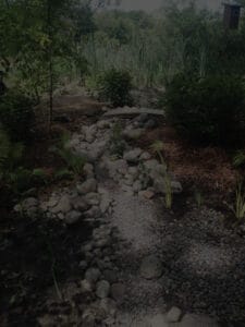 A darkened portrait of a landscaping design featuring a rocky dry creek bed with a custom with a small wooden bridge crossing it in front of a wetland.