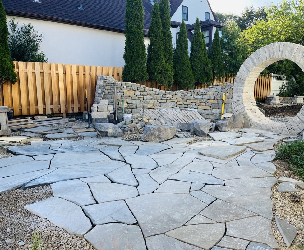 An in-progress flagstone patio, dry stacked rock wall and dramatic circular moon gate are key features of a rustic outdoor kitchen and beer garden.